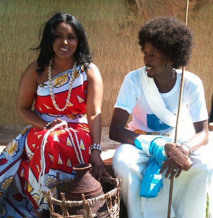 A Somali woman and man in traditional dress, she in a red patterned dirac with a cowrie necklace, he in white with a Somali flag scarf