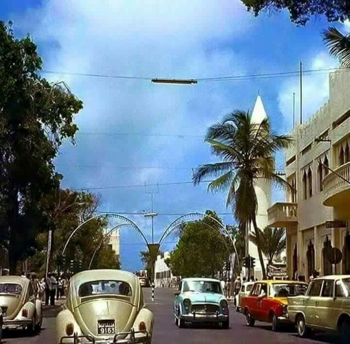 A busy Mogadishu street with classic cars, palm trees, a white minaret and metal arches